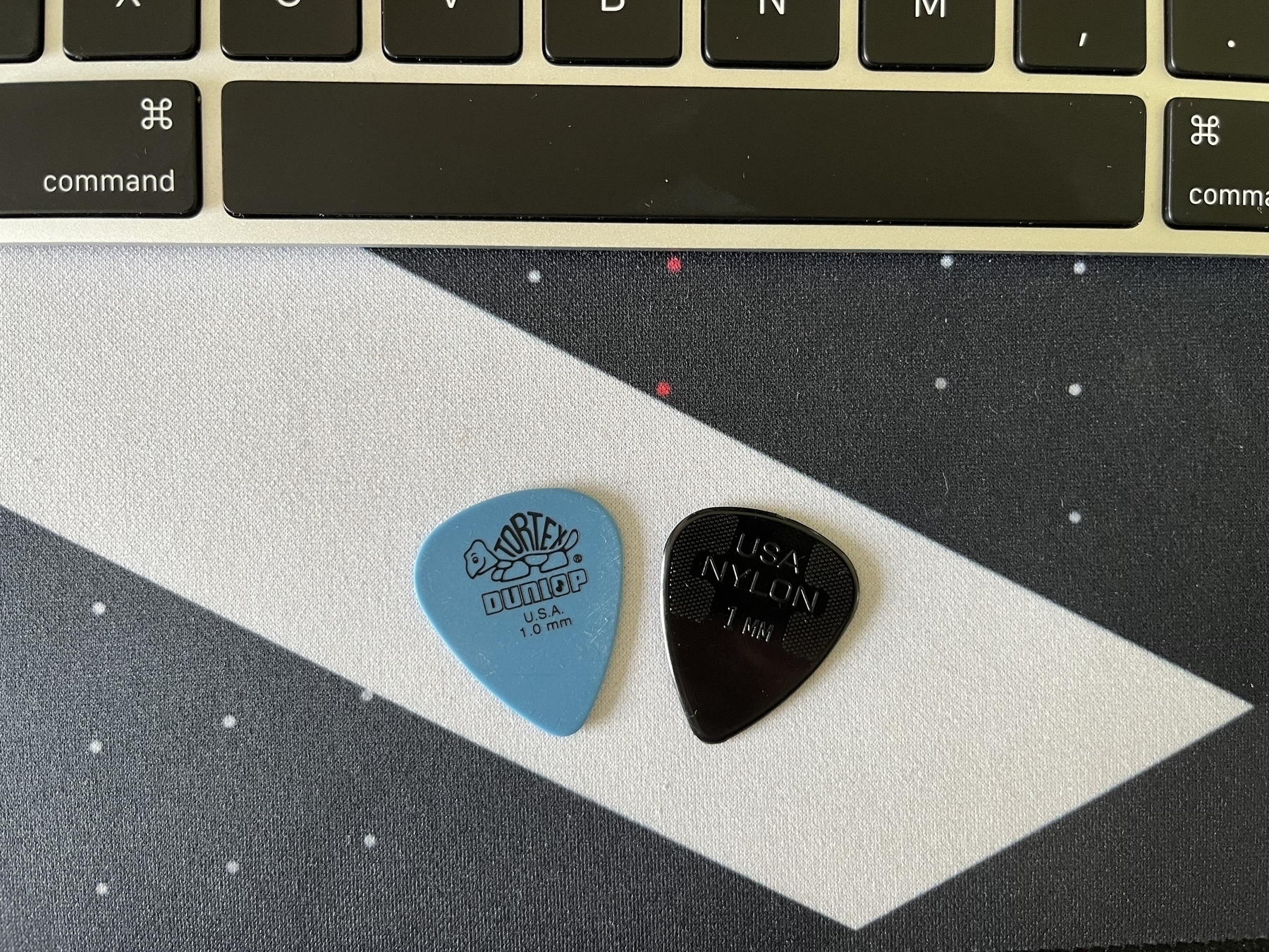 Two guitar picks: on the left is a blue Tortex 1mm pick, on the right is a black nylon 1mm pick.&10;&10;They are on an MKBHD desk mat. An Apple Magic Keyboard is visible near the top of the picture.